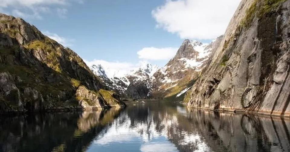 A still fjord between steep snowy mountains reflecting the sky in daylight